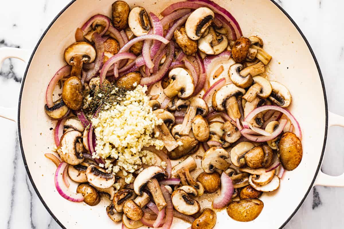 Large white sauté pan with sliced mushrooms, red onion, garlic, and dried herbs.