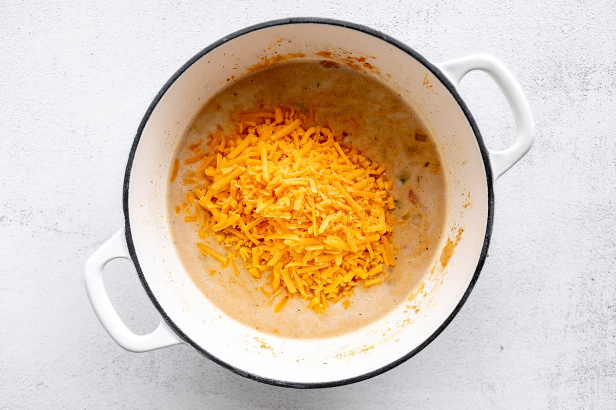 Shredded cheddar cheese added to a large white pot filled with creamy veggies.