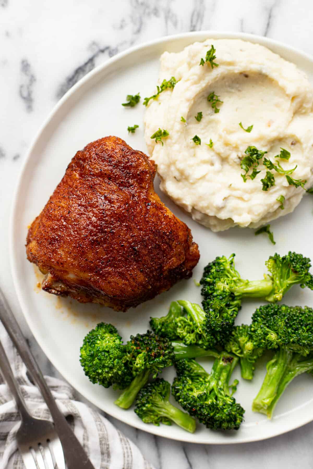 White plate with a smoked chicken thigh, mashed potatoes, and broccoli.