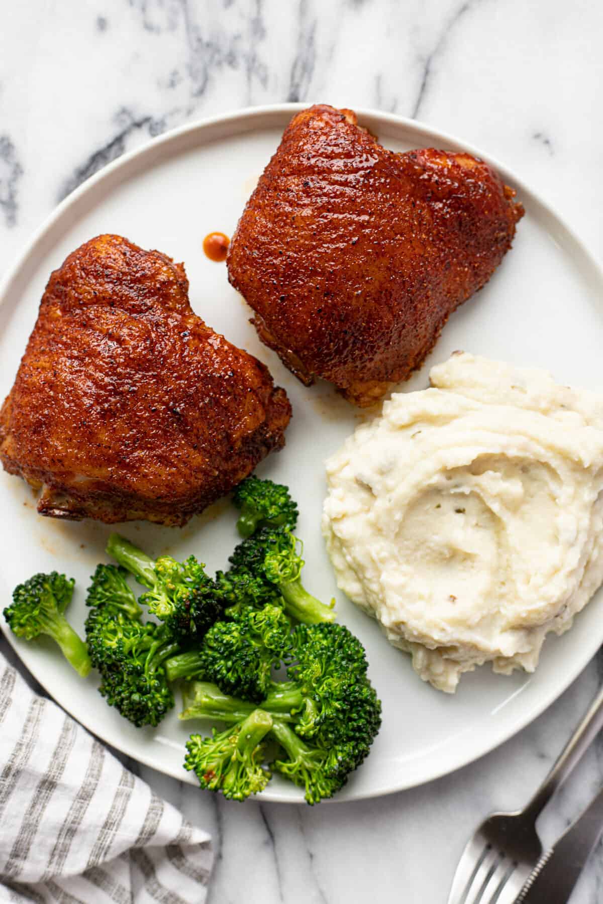 White plate with two smoked chicken thighs, mashed potatoes, and broccoli.