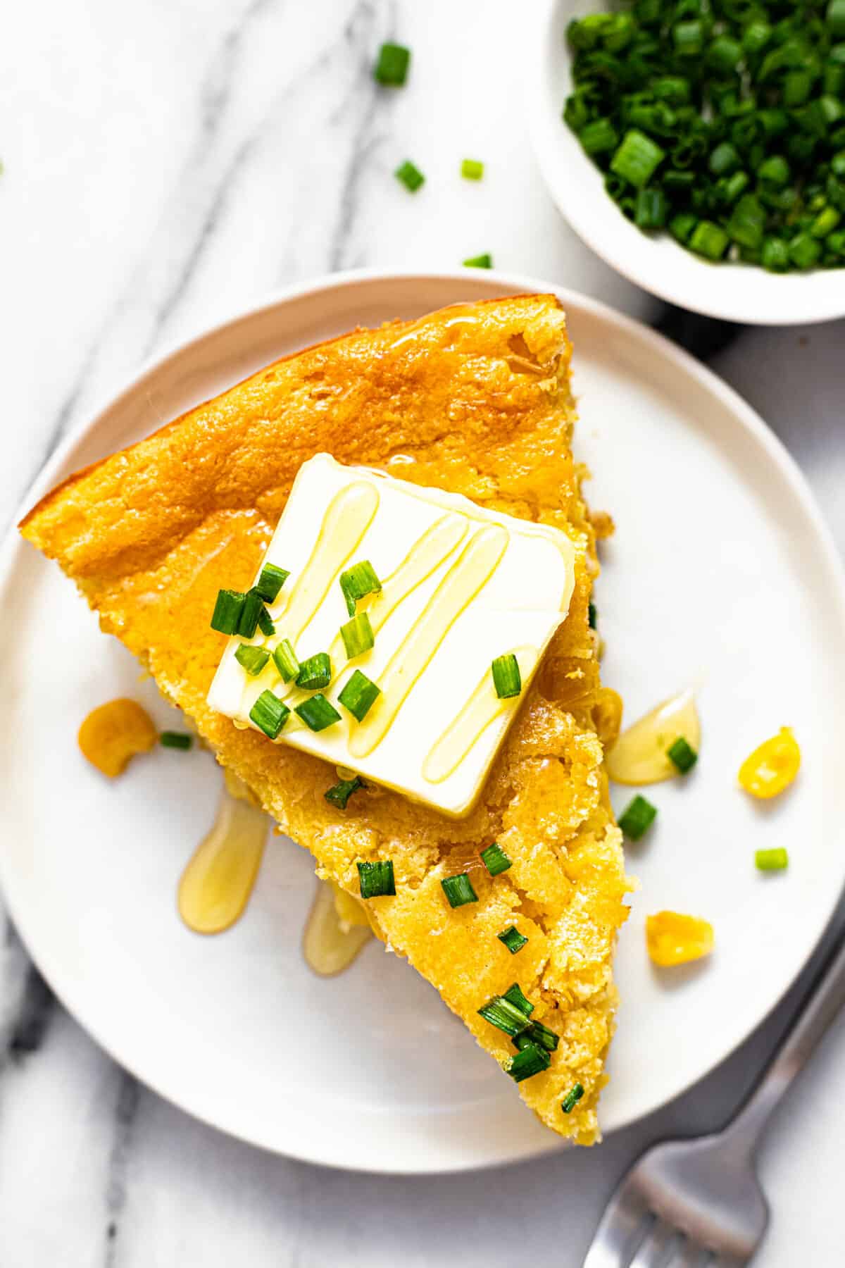 A slice of cornbread casserole on a white plate topped with butter, honey, and chives.