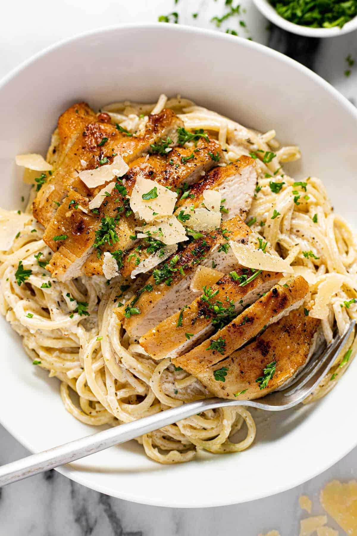 Large white bowl filled with creamy spaghetti and sliced lemon chicken garnished with parsley.