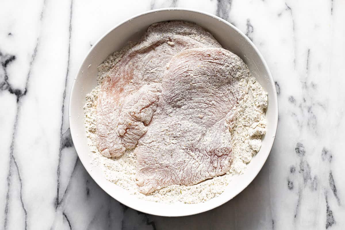 Chicken breast dredged in a white bowl filled with flour and seasonings. 