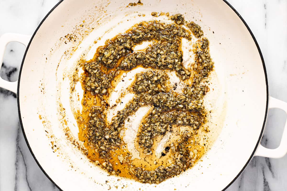 Sauteed garlic, herbs, and spices in olive oil in a large white pan. 