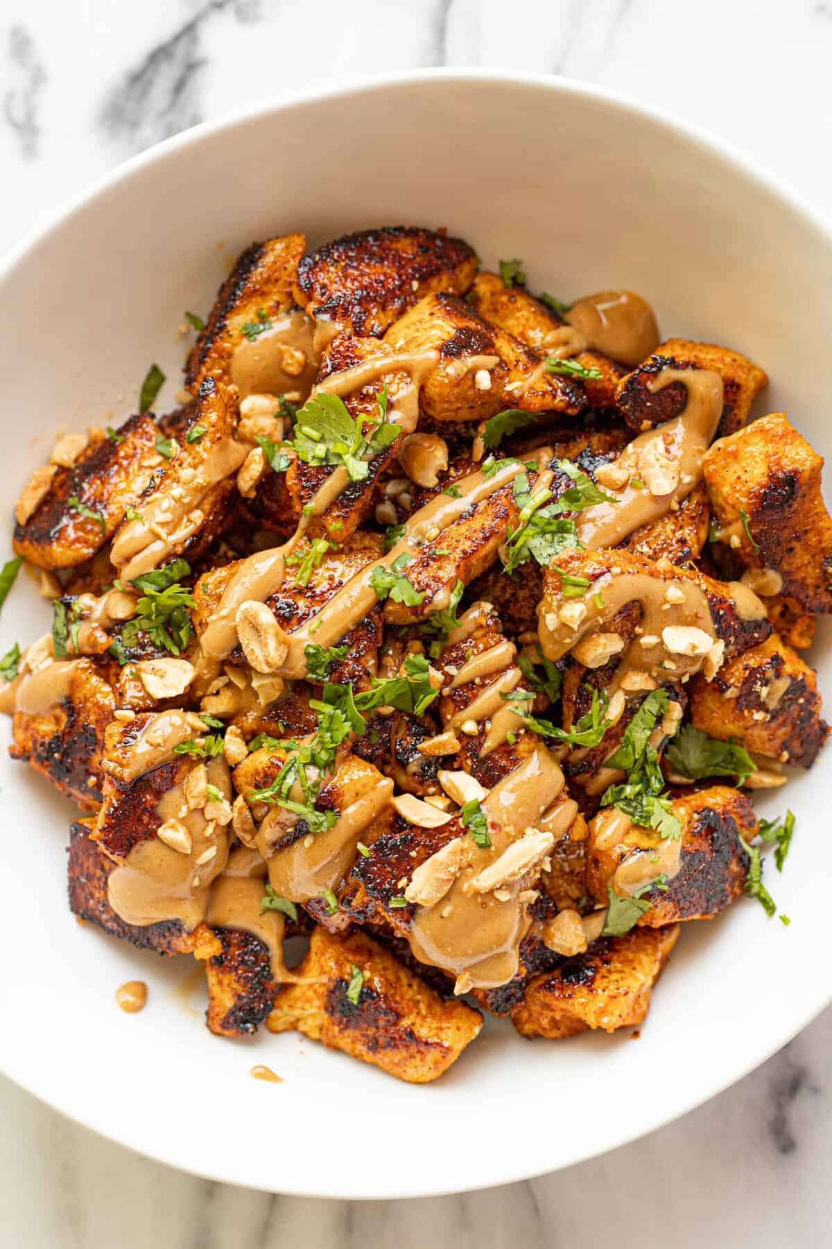 Sauteed chicken in a white bowl garnished with peanut sauce, cilantro, and peanuts. 