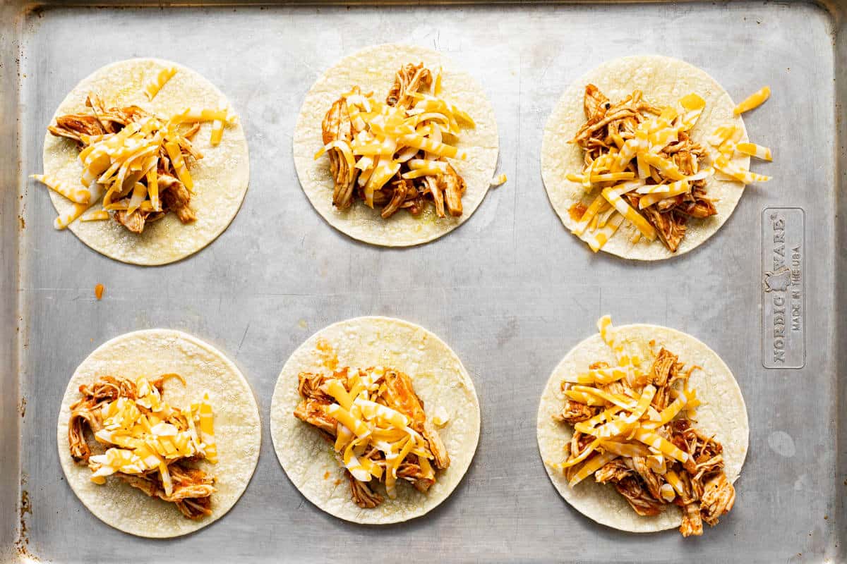 Small corn tortillas topped with shredded chicken and shredded cheese on a metal baking sheet.