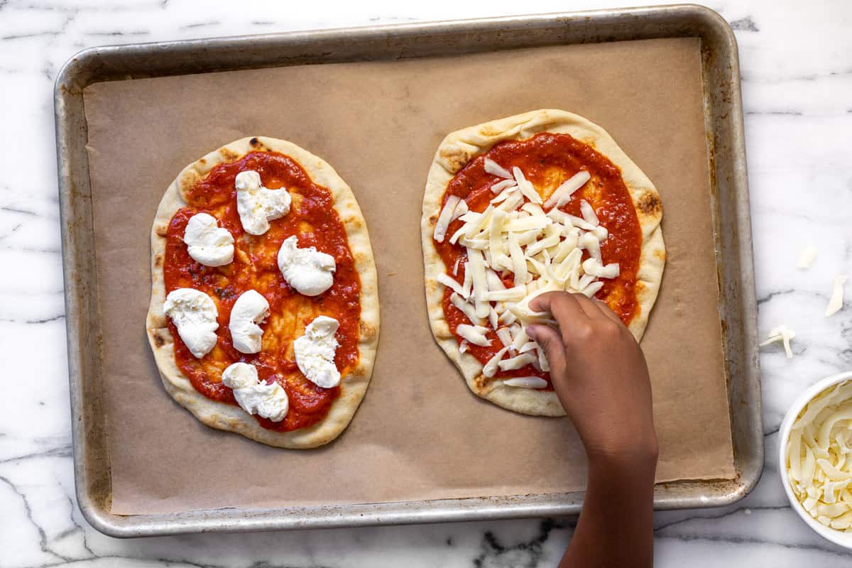 Small hand adding cheese to two naan pizzas on a parchment lined baking sheet.