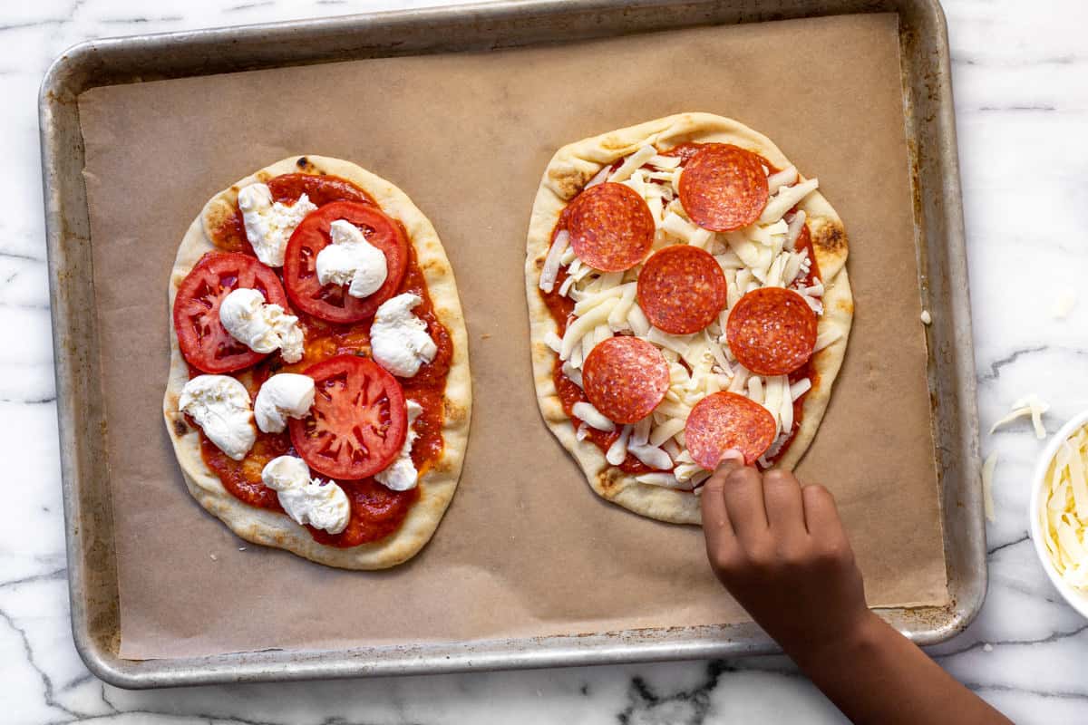 Small hand adding pepperoni to a naan pizza on a parchment lined baking sheet.