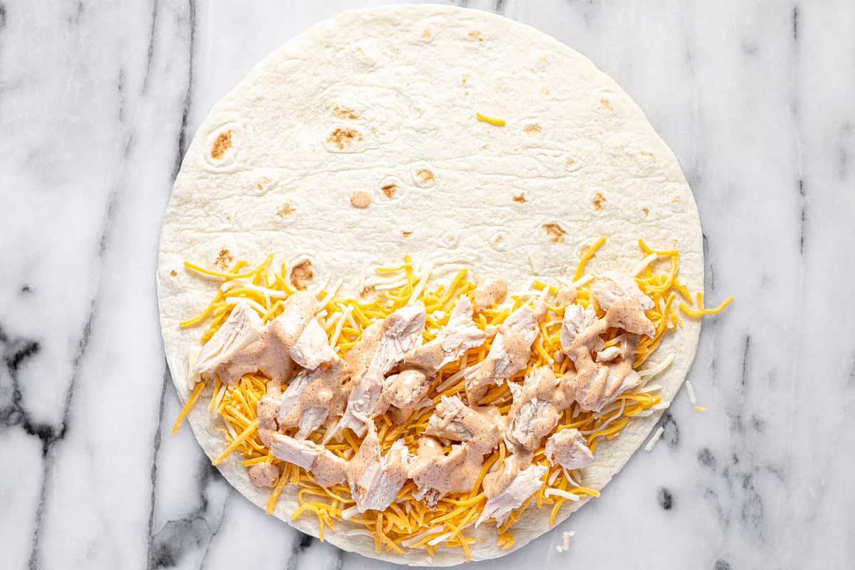 A flour tortilla with shredded cheese, diced grilled chicken, and creamy quesadilla sauce on it.