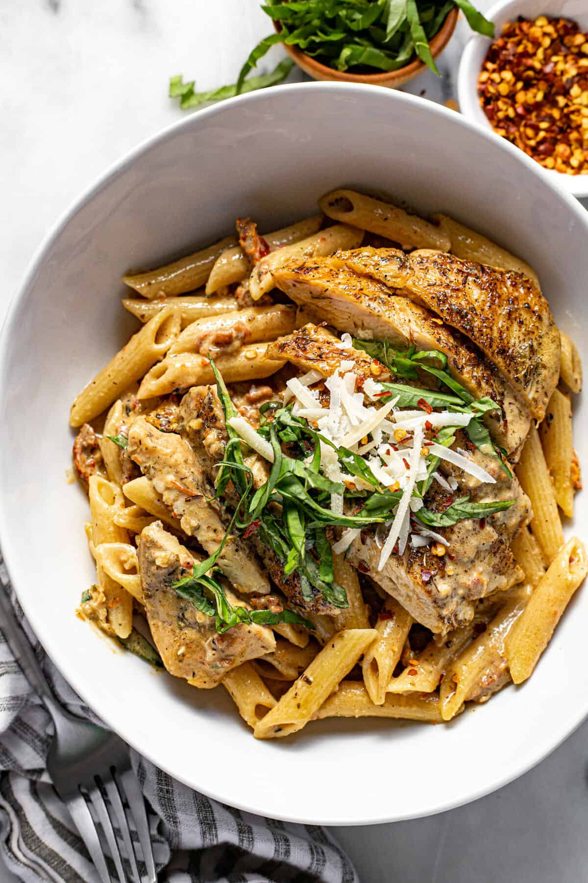 White bowl of pasta topped with sliced marry me chicken, fresh basil, and Parmesan cheese.