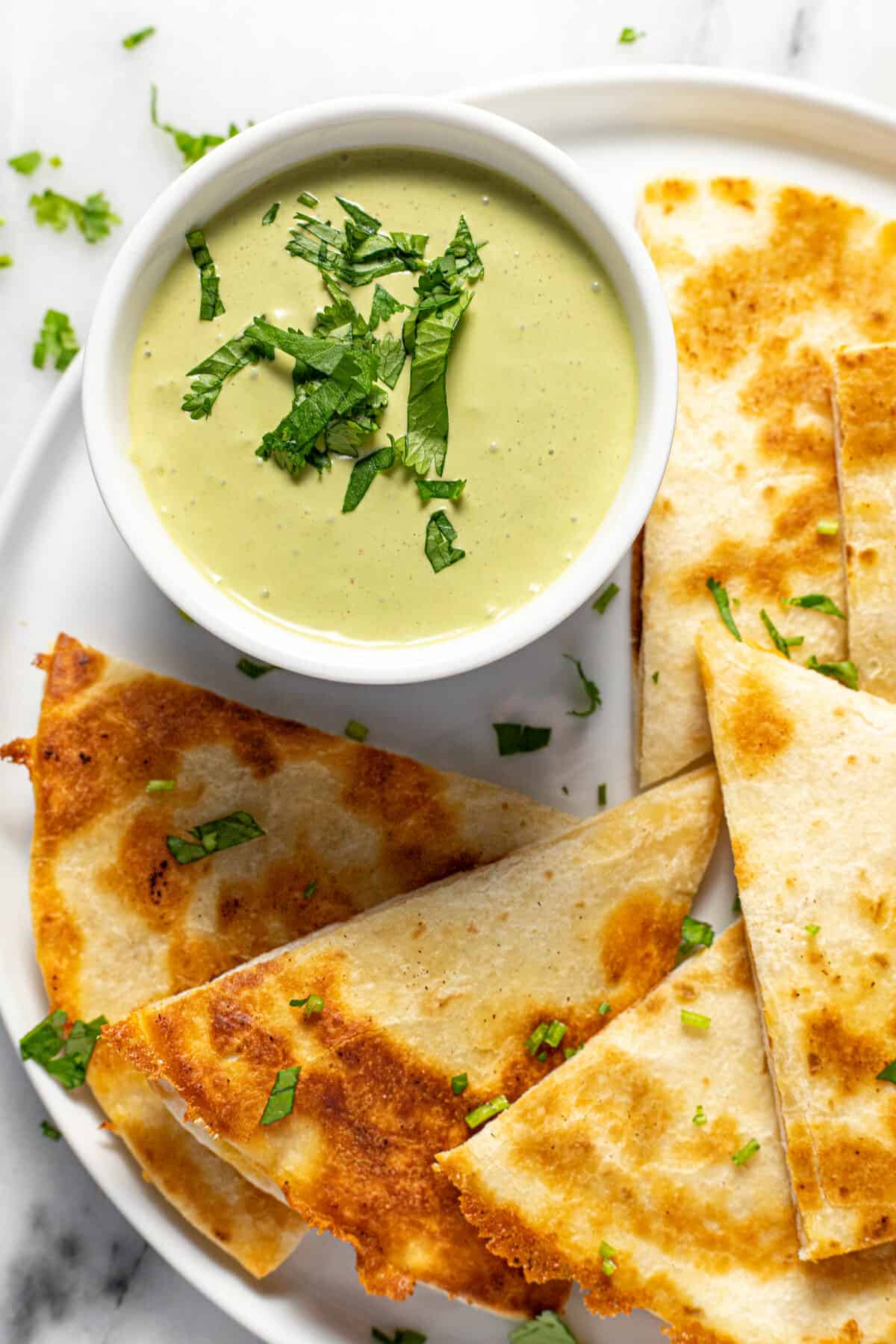 Small white bowl filled with cilantro lime sauce on a plate with quesadillas.