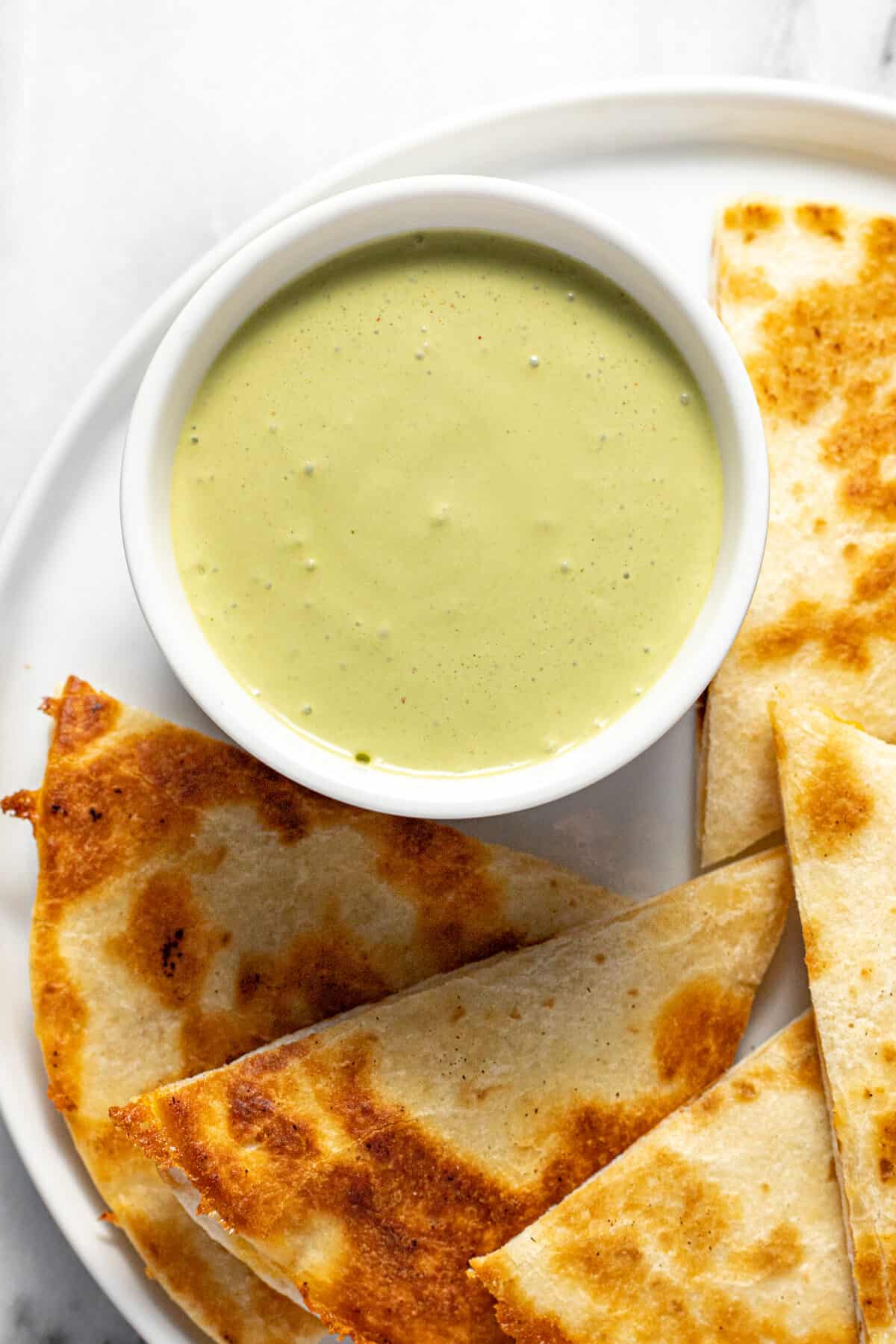 Small white bowl filled with cilantro lime sauce on a plate with quesadillas.