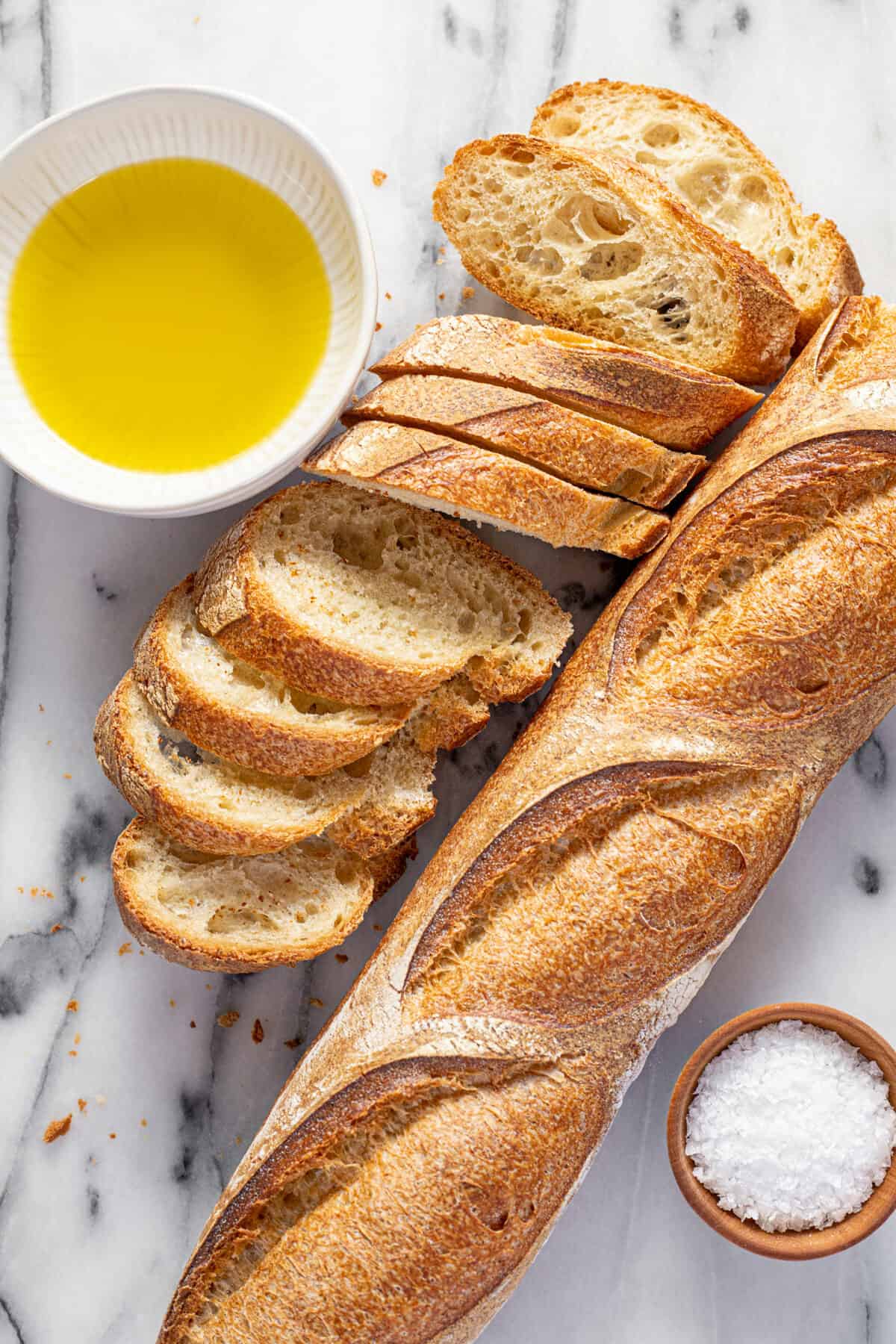 White marble counter top with sliced baguette, a small bowl of salt, and a bowl of olive oil. 