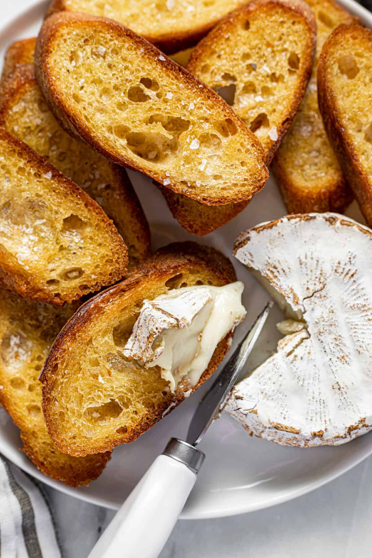A white plate filled with crostini with a small wheel of brie being sliced with a cheese knife.