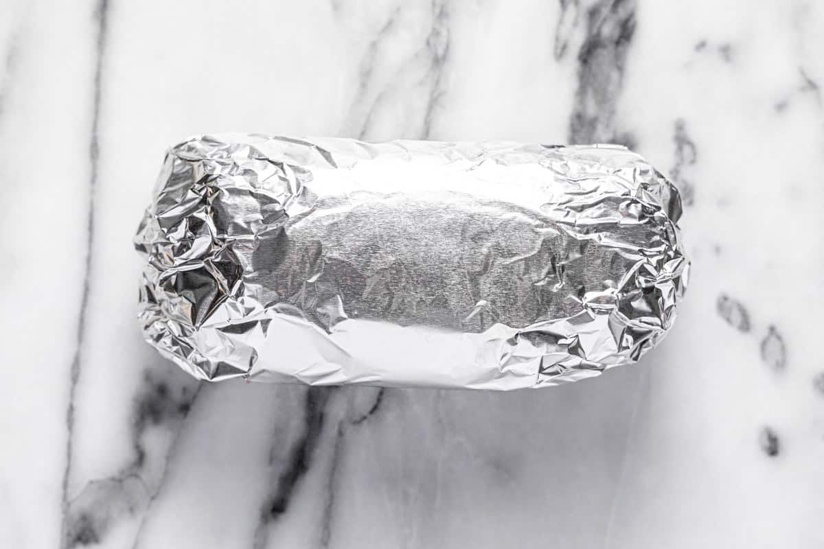 A California burrito wrapped in tin foil on a white marble counter top.