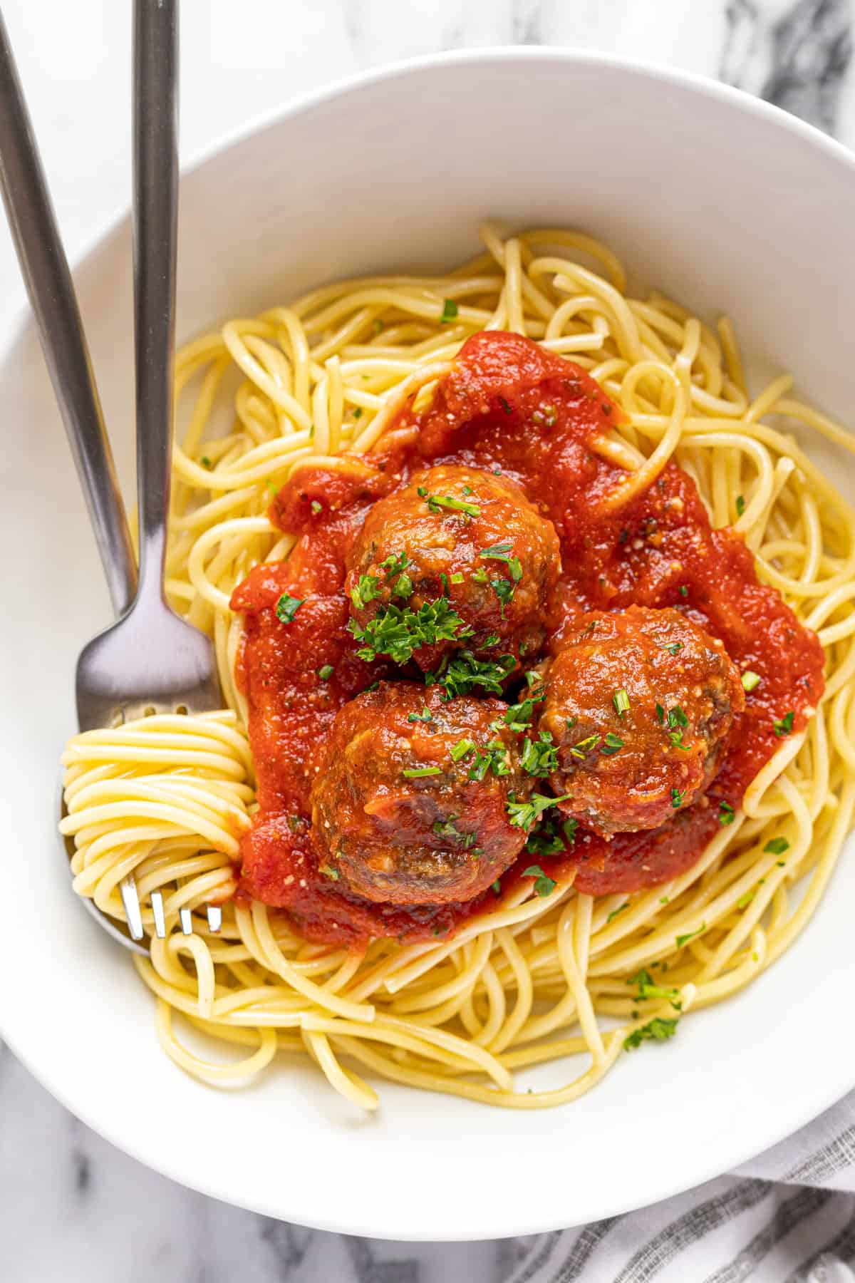 White bowl filled with spaghetti and homemade meatballs with marinara sauce and parsley.