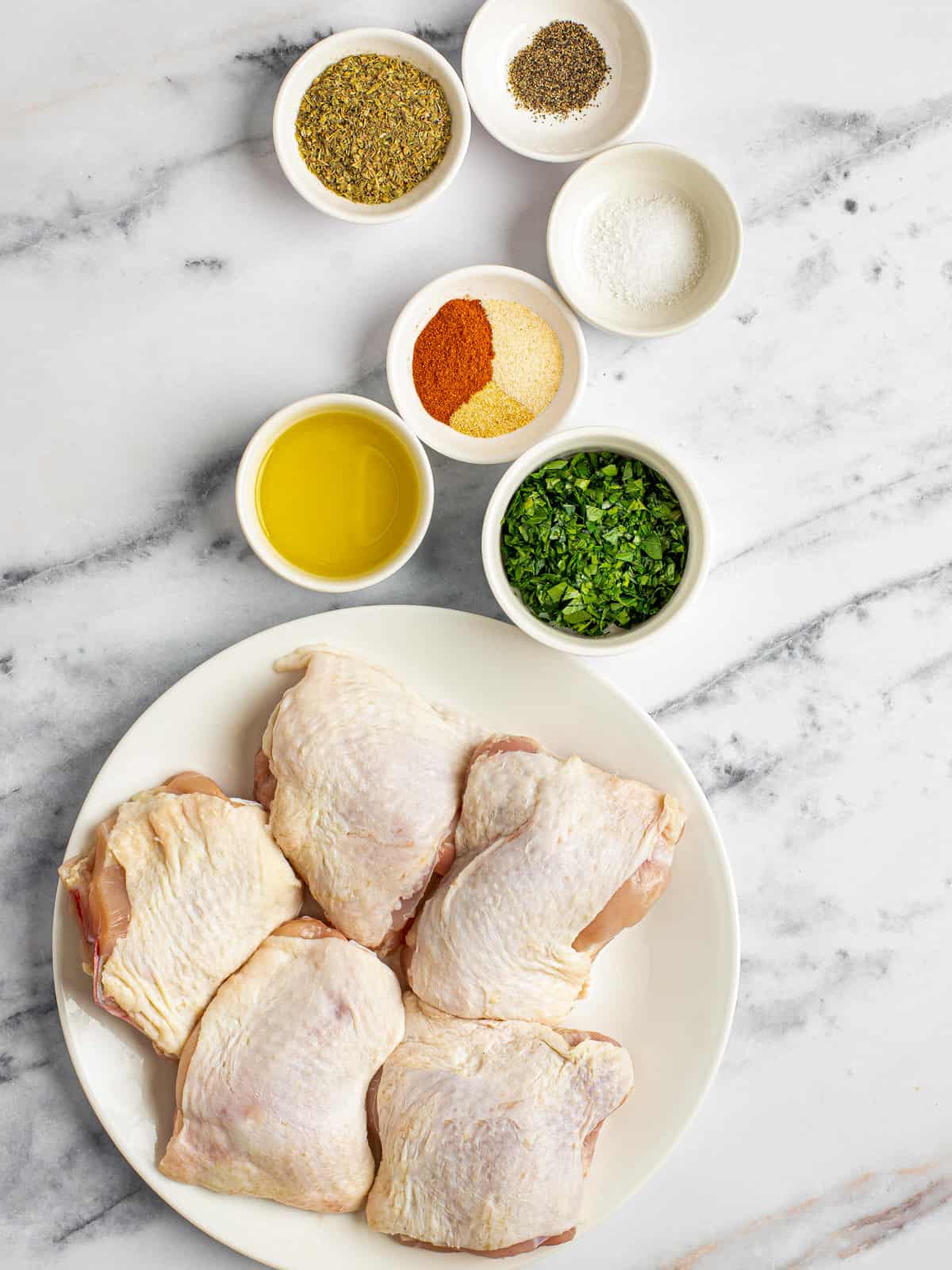 White marble counter top with bowls of ingredients to make juicy baked chicken thighs.
