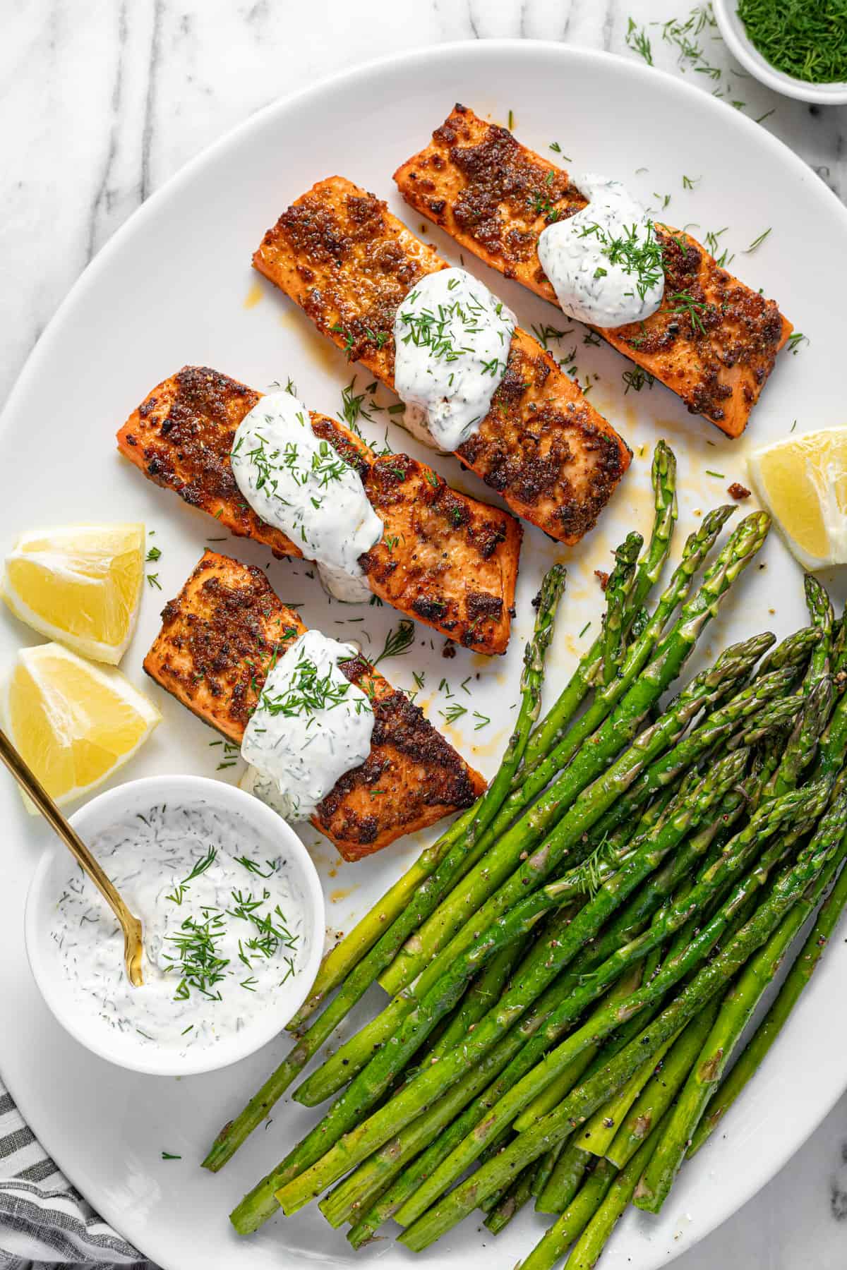 White serving platter with 4 fillets of salmon drizzled with dill sauce with sauteed asparagus.