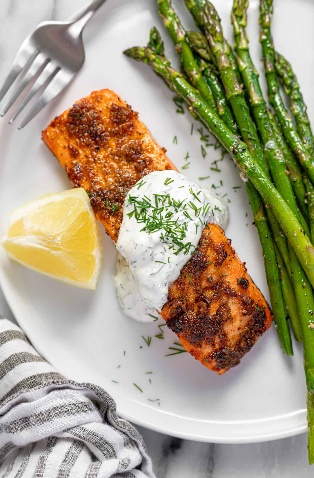White plate with 1 fillet of salmon drizzled with dill sauce, sauteed asparagus, and a wedge of lemon.