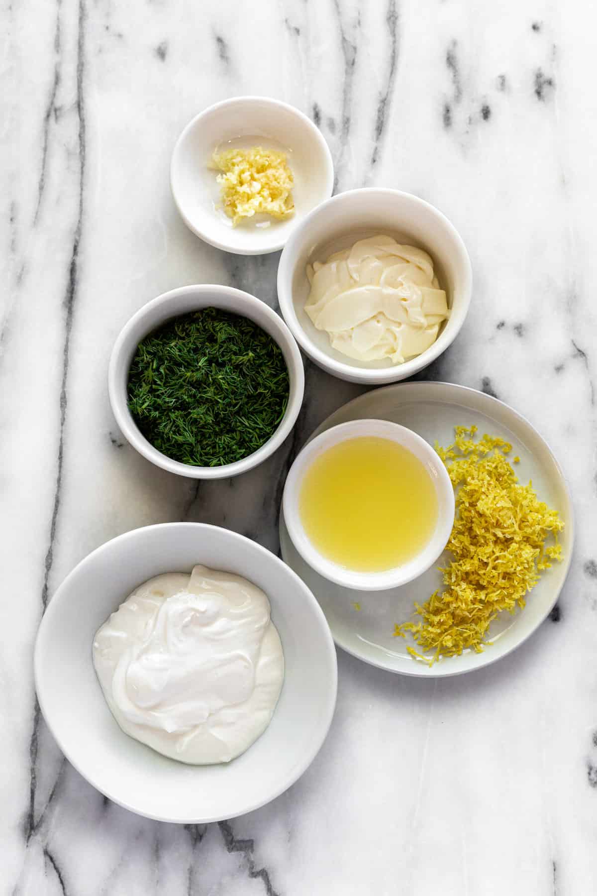White marble countertop with bowls of ingredients to make creamy dill sauce for salmon. 
