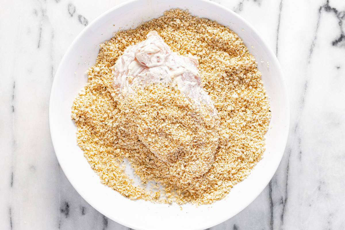 A mayo coated chicken breast being tossed in a bowl of breadcrumbs and Parmesan cheese. 