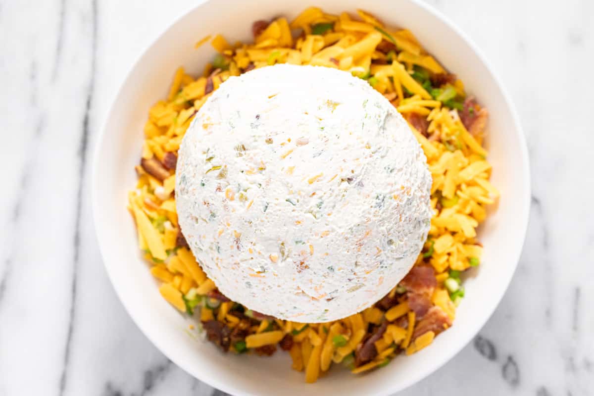 A big cheese ball in a bowl of shredded cheese, bacon, and green onion. 