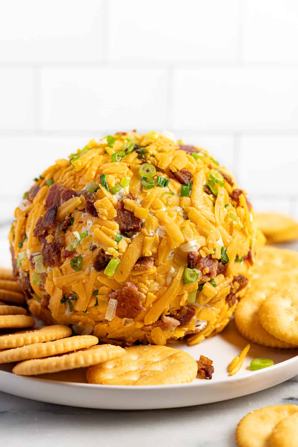 Big round cheese ball coated in shredded cheese, bacon, and green onion on a round plate with crackers. 