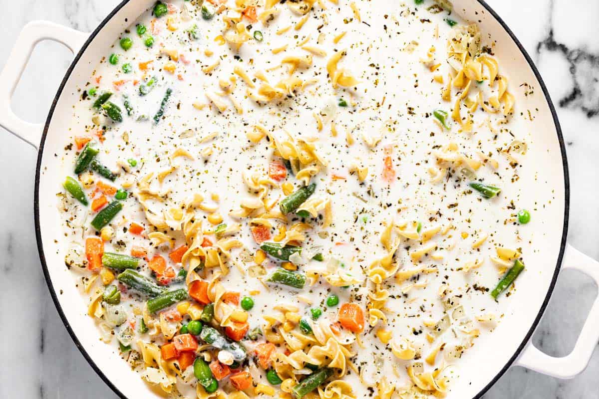 Large saute pan filled with uncooked pasta, heavy cream, broth, and frozen veggies. 