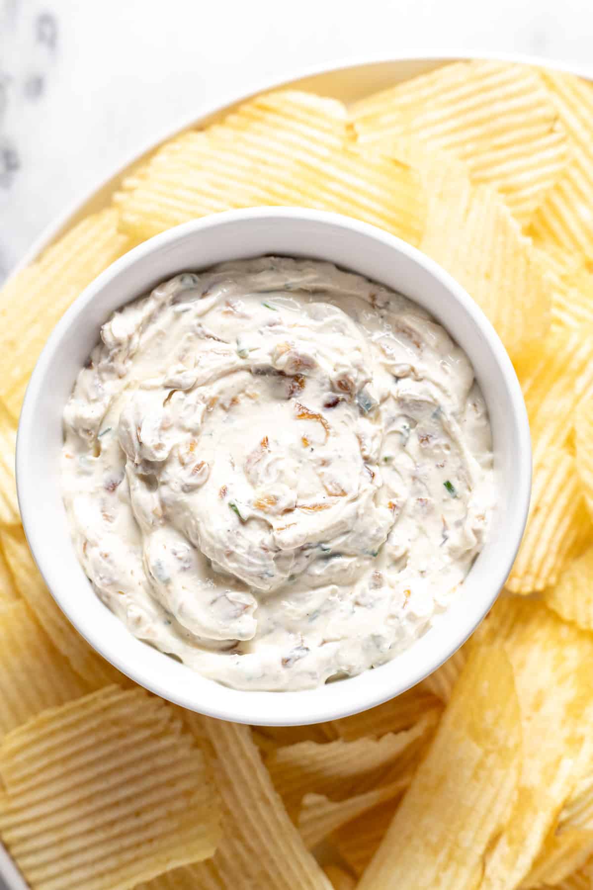 Small bowl of French onion dip on a plate of potato chips.