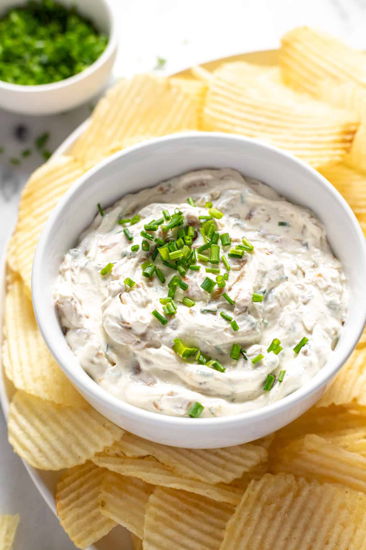 Small bowl of French onion dip on a plate of potato chips garnished with chives. 
