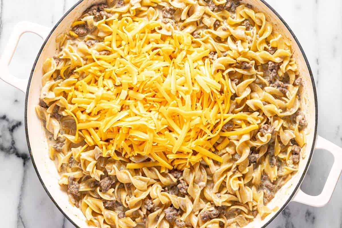 Shredded cheese being added to a large pan of creamy beef and noodles. 