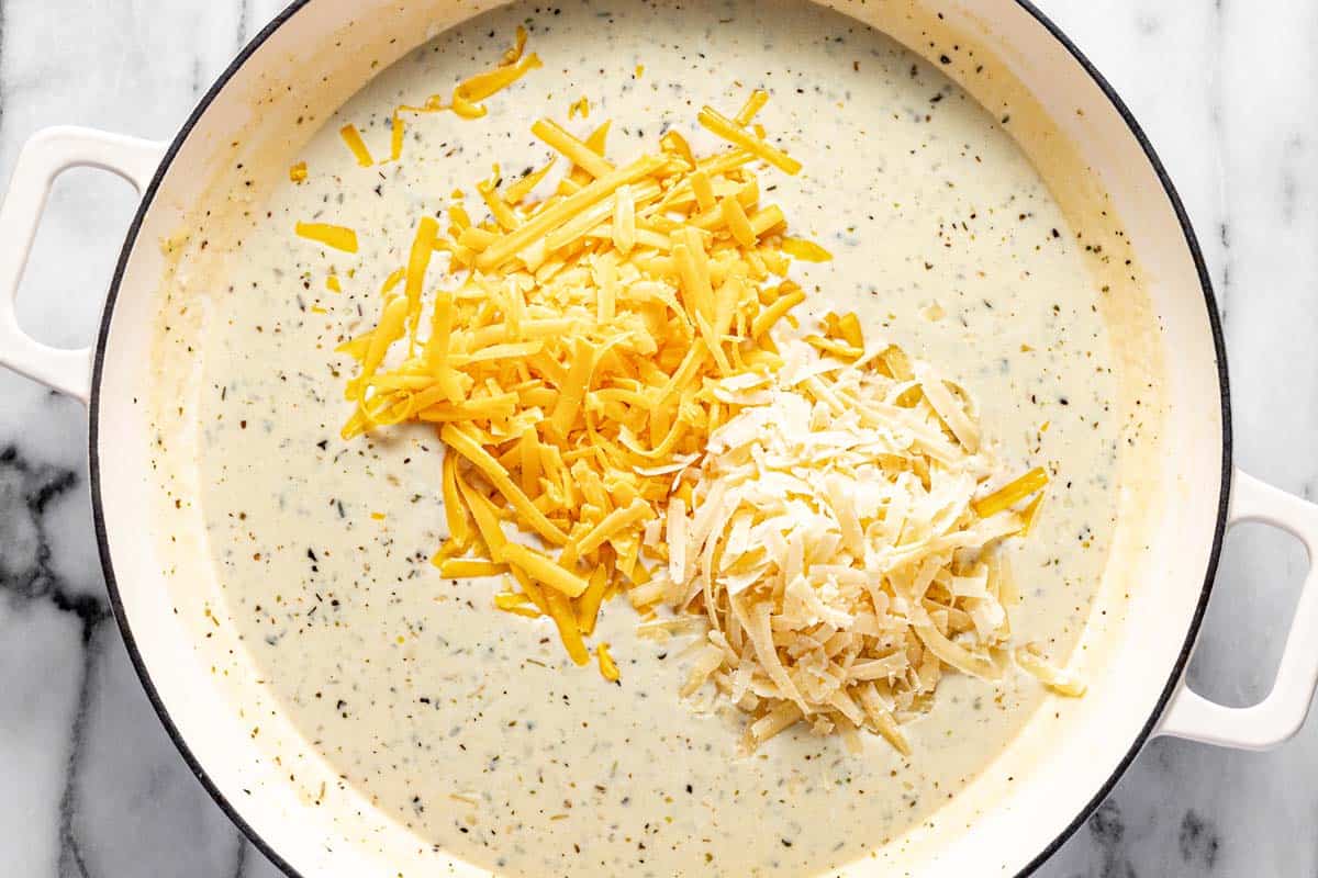 Shredded cheese being added to a creamy sauce in a large pan. 