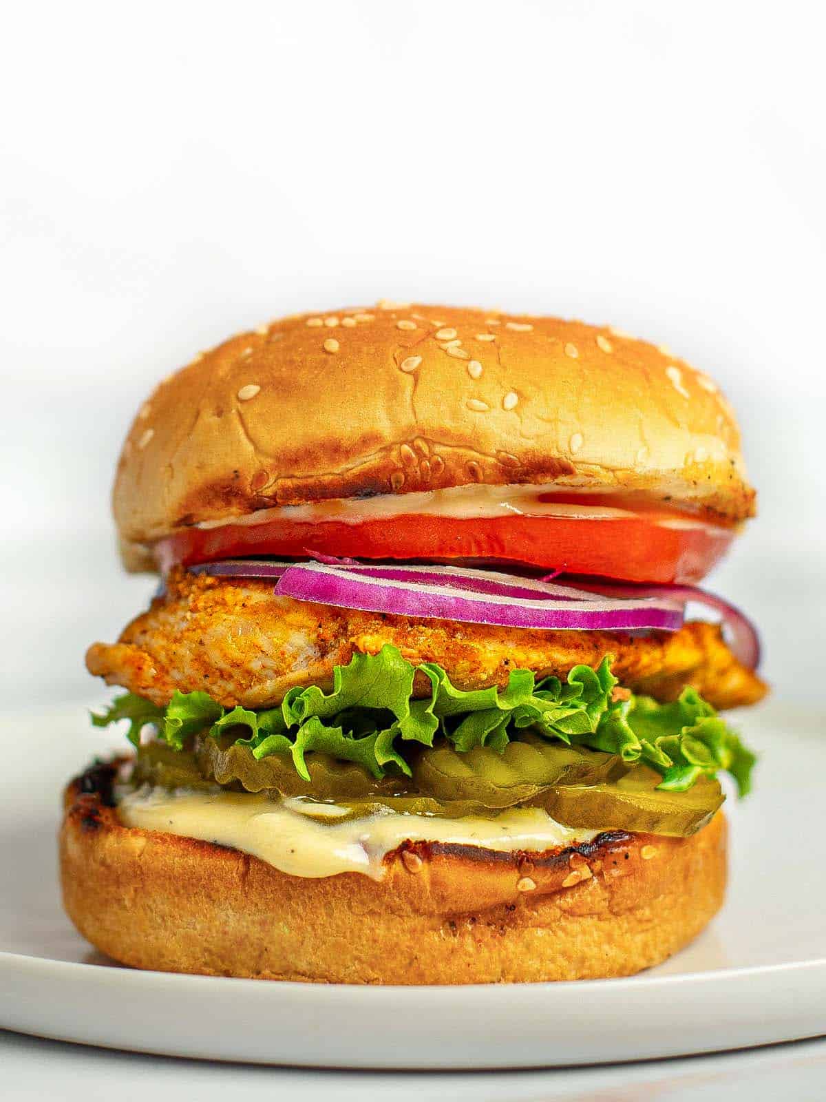 Close up shot of a grilled chicken sandwich stacked with lettuce, tomato, onion, pickles, and honey mustard dressing. 