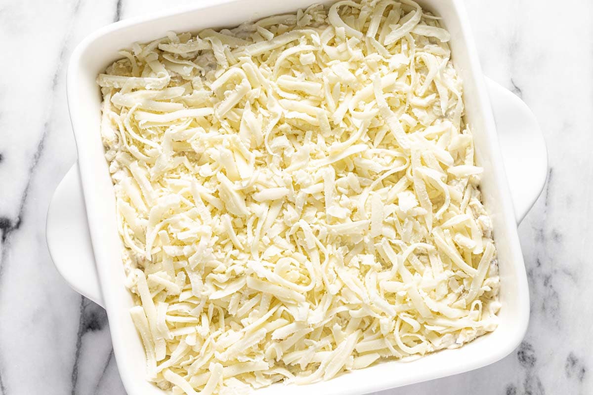 Shredded cheese on top of artichoke dip in a square baking dish. 