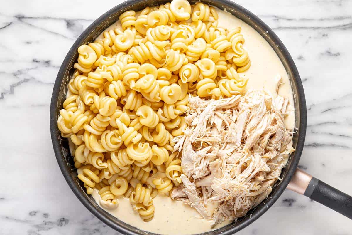 Pasta and shredded chicken added to a large pan of creamy cheese sauce. 