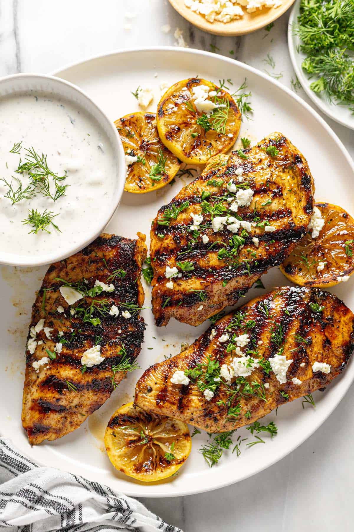 White plate with grilled Greek chicken, sliced lemons, feta, and fresh herbs.