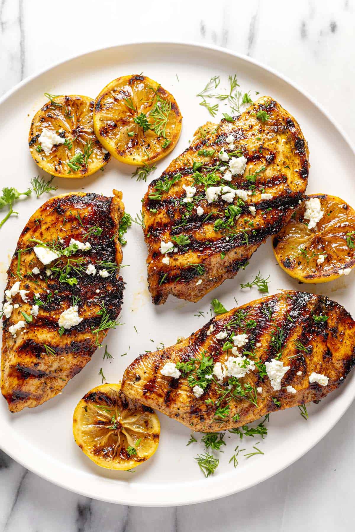 Large plate with grilled Greek chicken, lemon slices, feta cheese, and fresh herbs. 