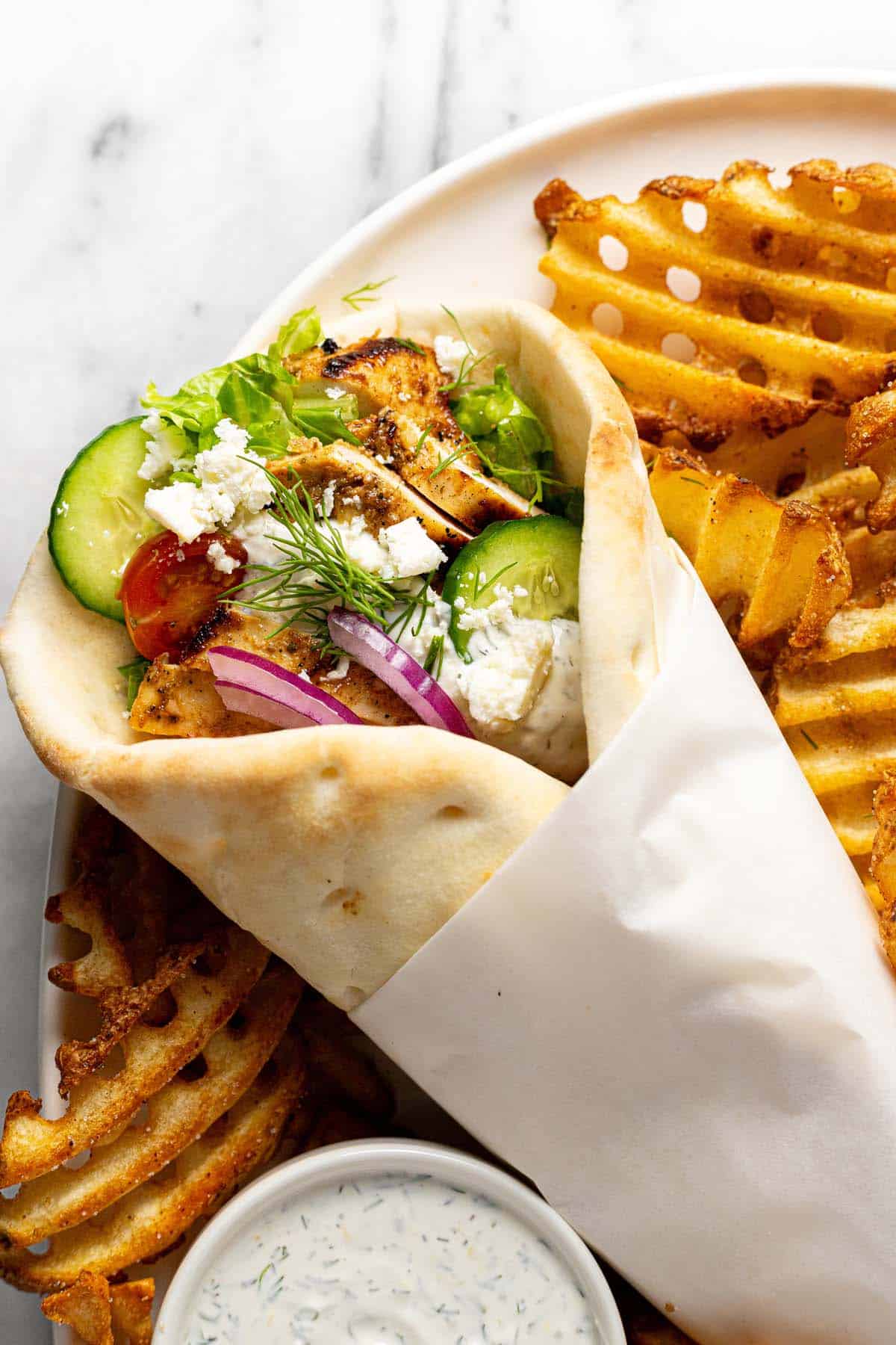 Close up shot of a Greek chicken gyro on a plate with waffle fries. 