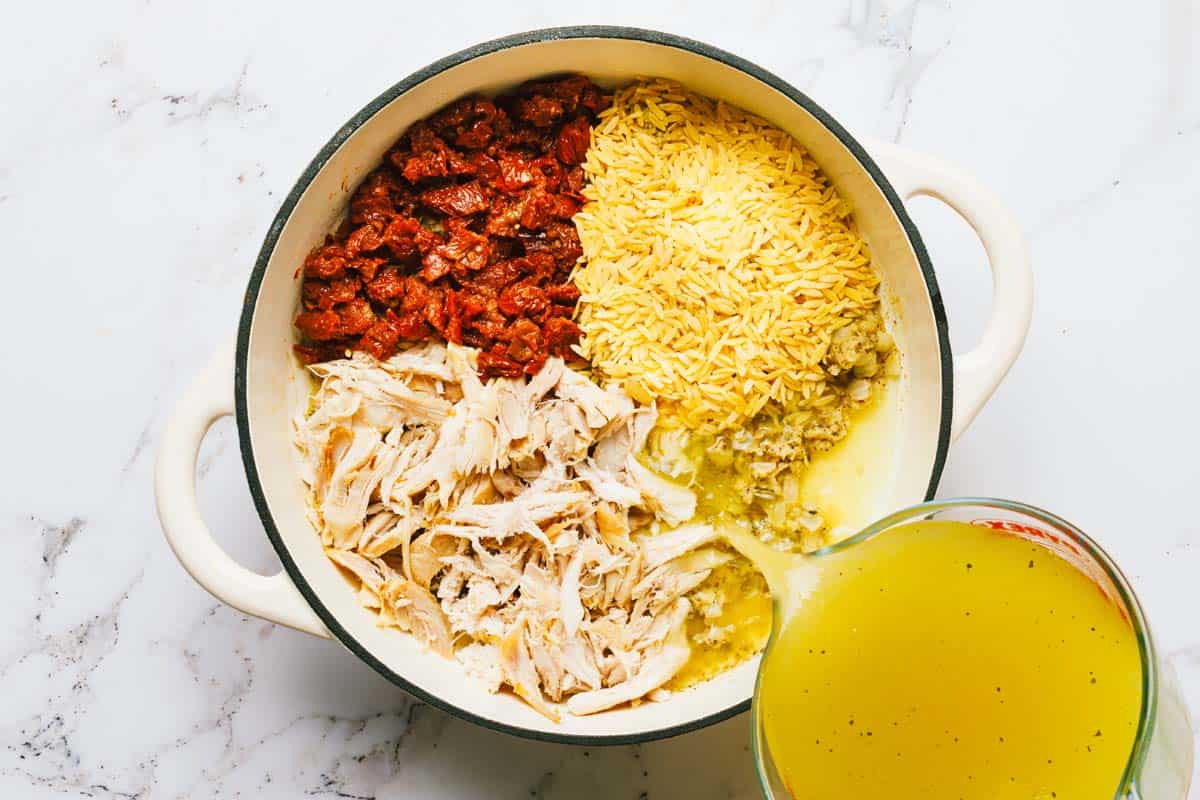 Sun-dried tomatoes, orzo, and shredded chicken in a pot with broth being added. 