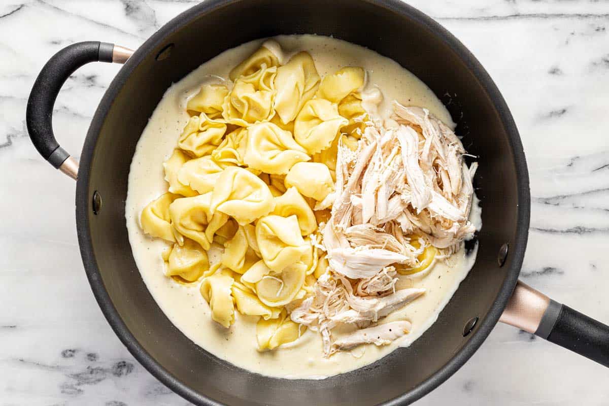 Tortellini and shredded chicken added to a pan of alfredo sauce. 