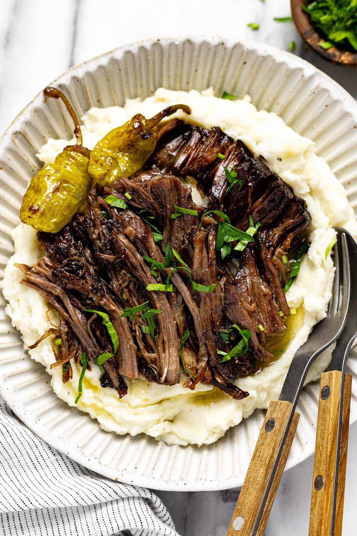 Bowl of mashed potatoes topped with shredded Mississippi pot roast garnished with chopped parsley. 