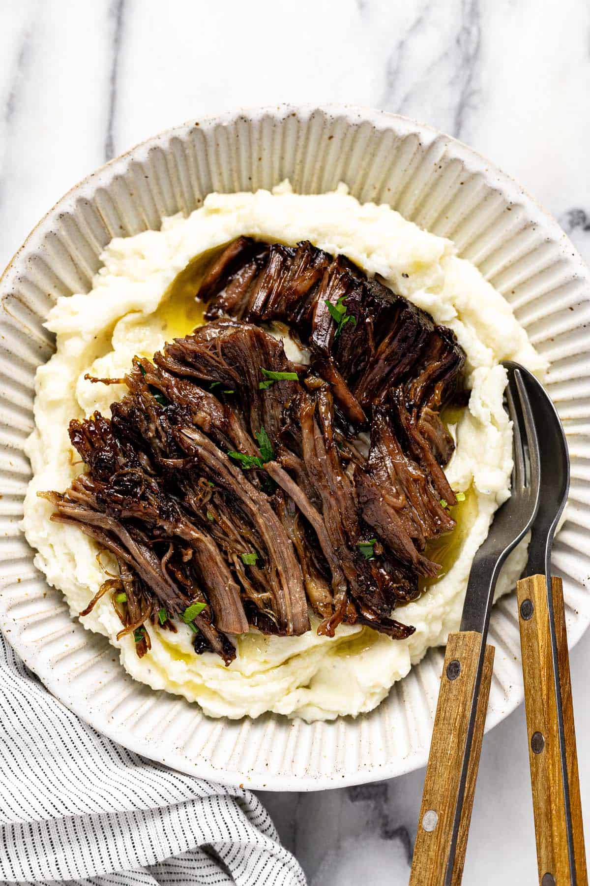 Bowl of mashed potatoes topped with shredded Mississippi pot roast garnished with chopped parsley. 