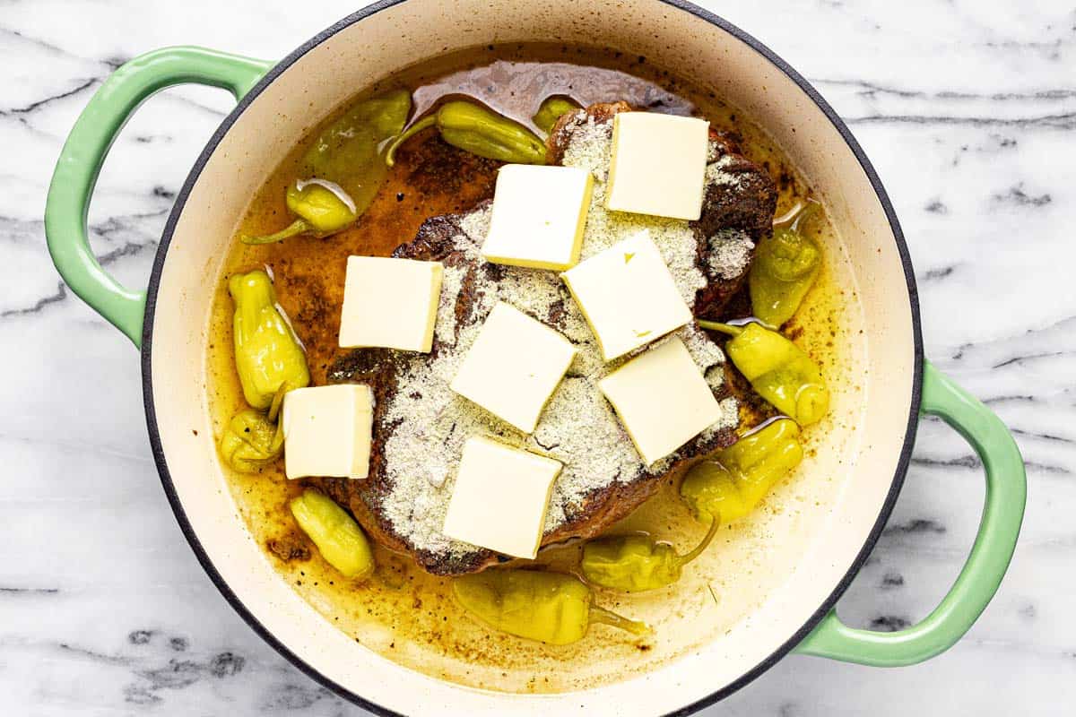 Seared roast in a pan with seasonings, sliced butter, and pepperoncini. 