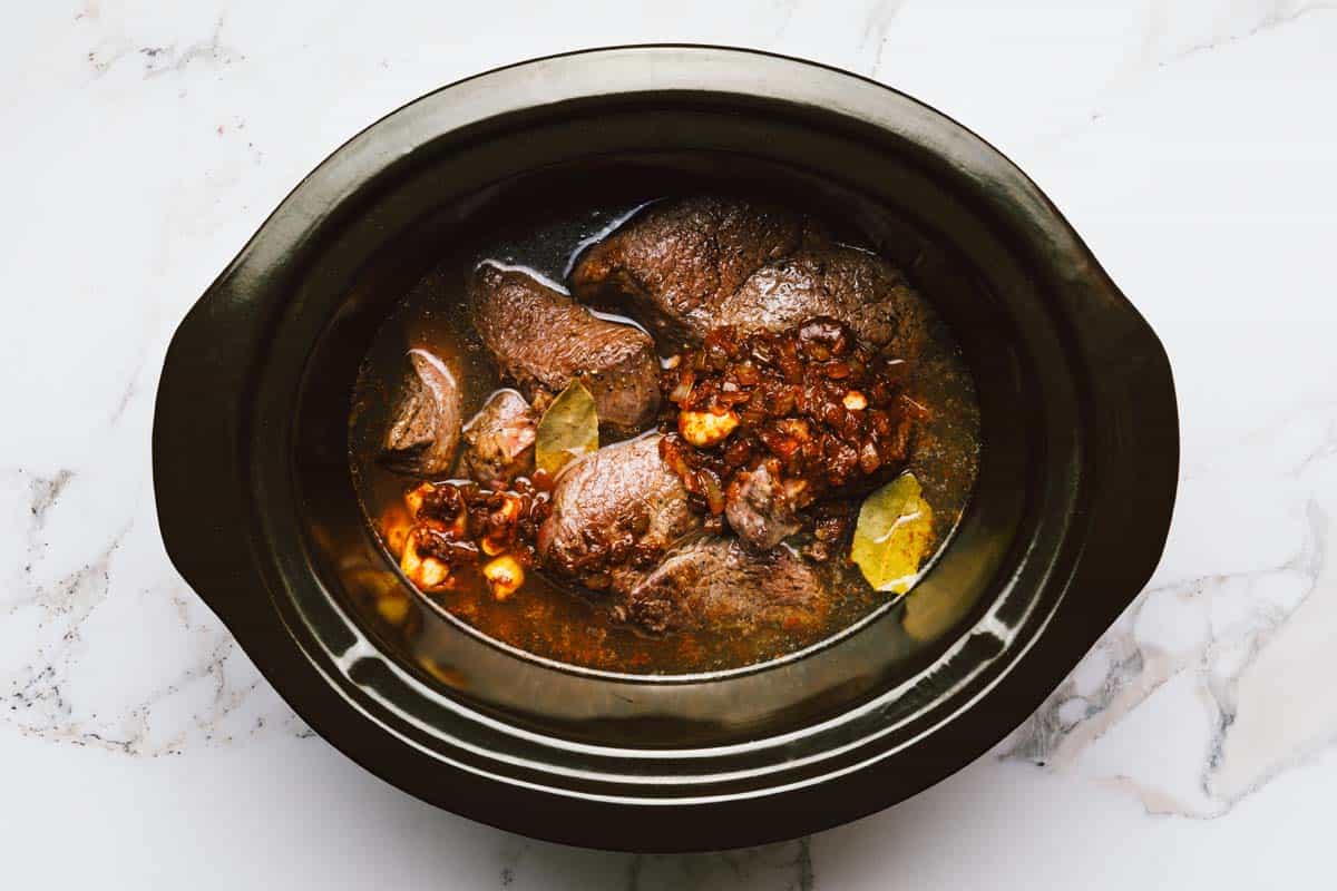 Ingredients to make shredded beef tacos in a slow cooker insert. 