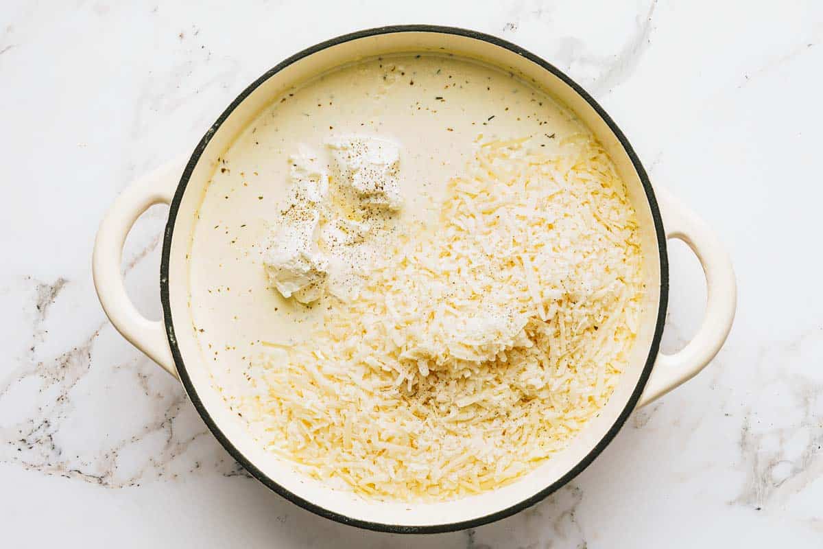 Cream cheese and shredded cheese being added to a pan of alfredo sauce. 