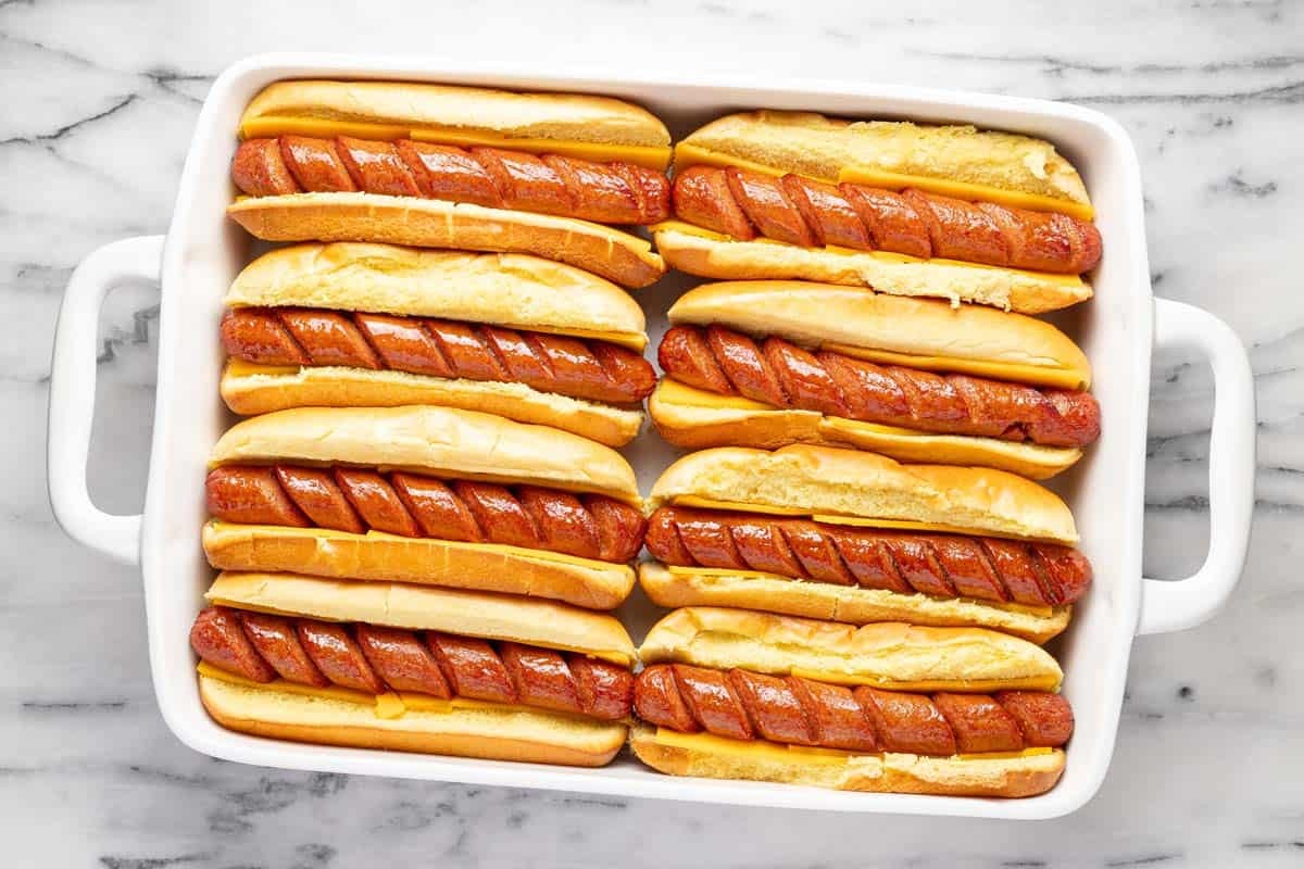 Baking dish filled with hot dog buns, sliced cheese, and cooked hot dogs. 