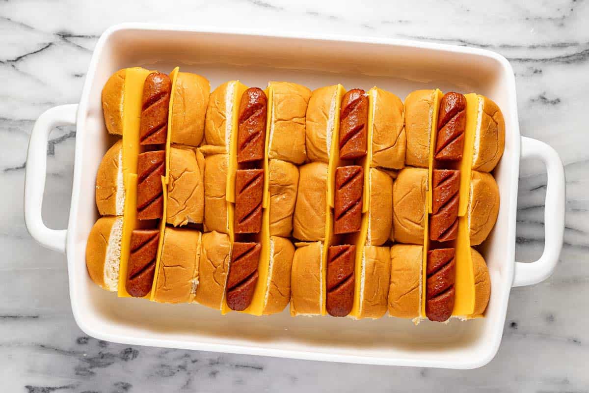 Baking dish with Hawaiian rolls, sliced cheese, and hot dogs in it. 