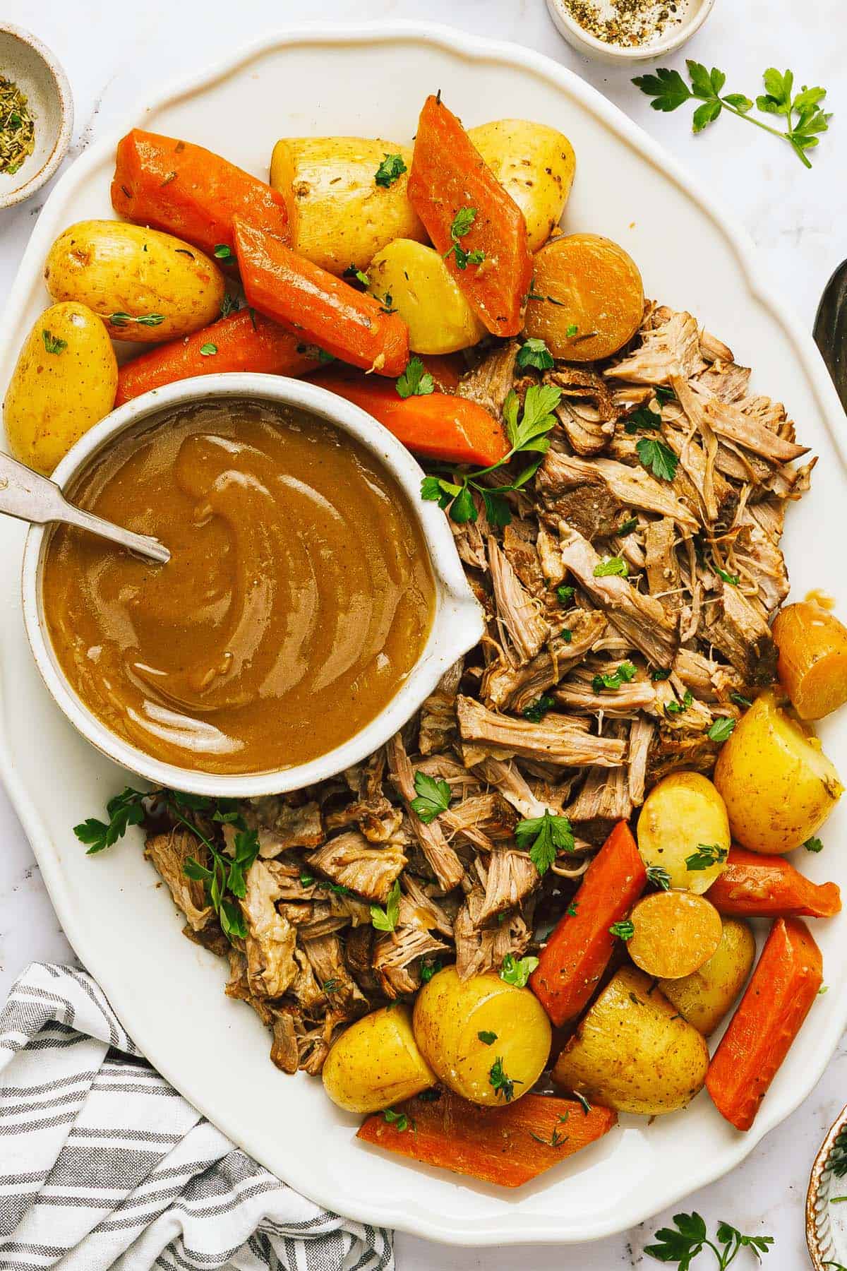 Large platter with a shredded pork roast, veggies, and a bowl of gravy on it. 