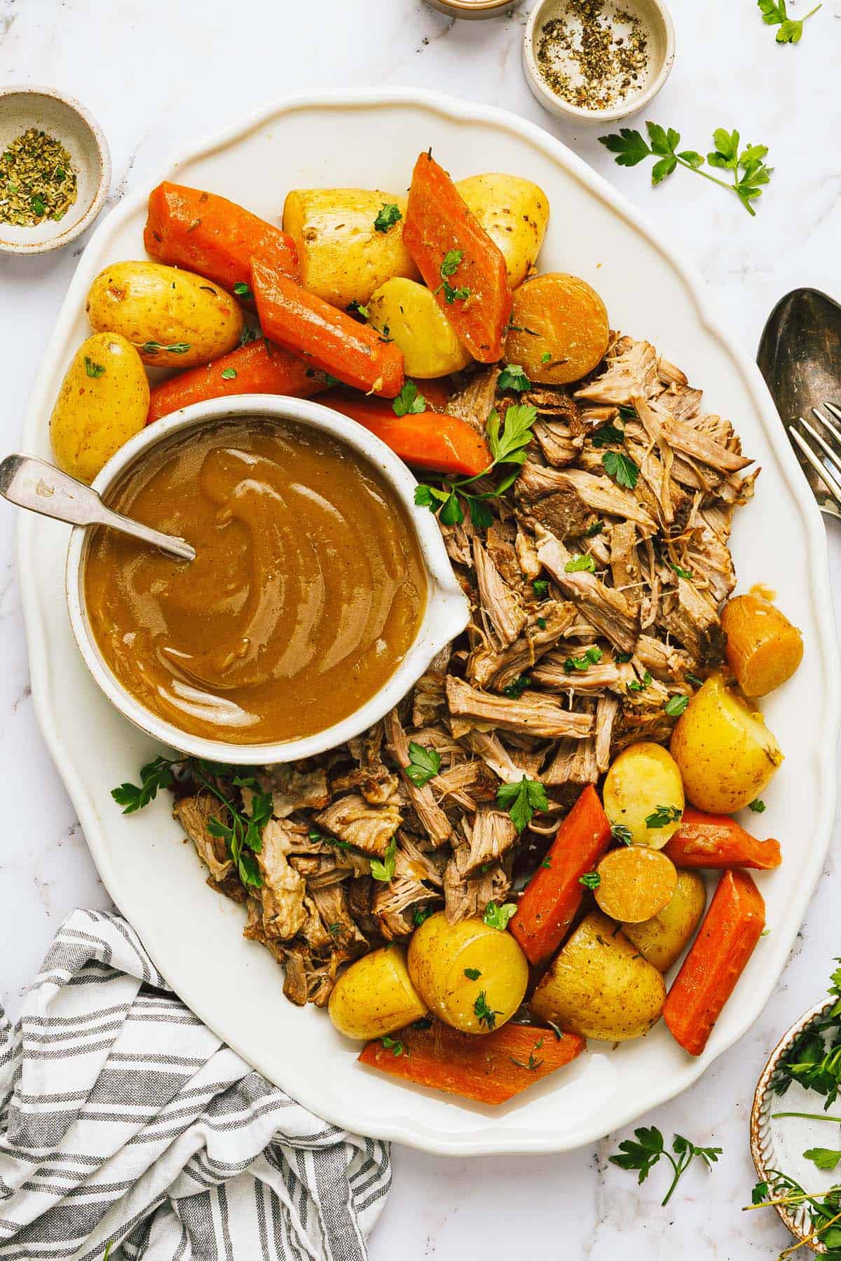 Large platter with a shredded pork roast, veggies, and a bowl of gravy on it. 