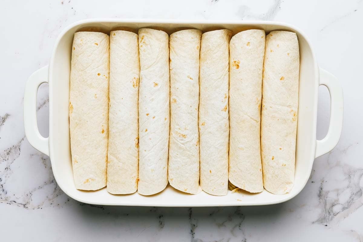 White chicken enchiladas rolled and seam side down in a baking dish. 